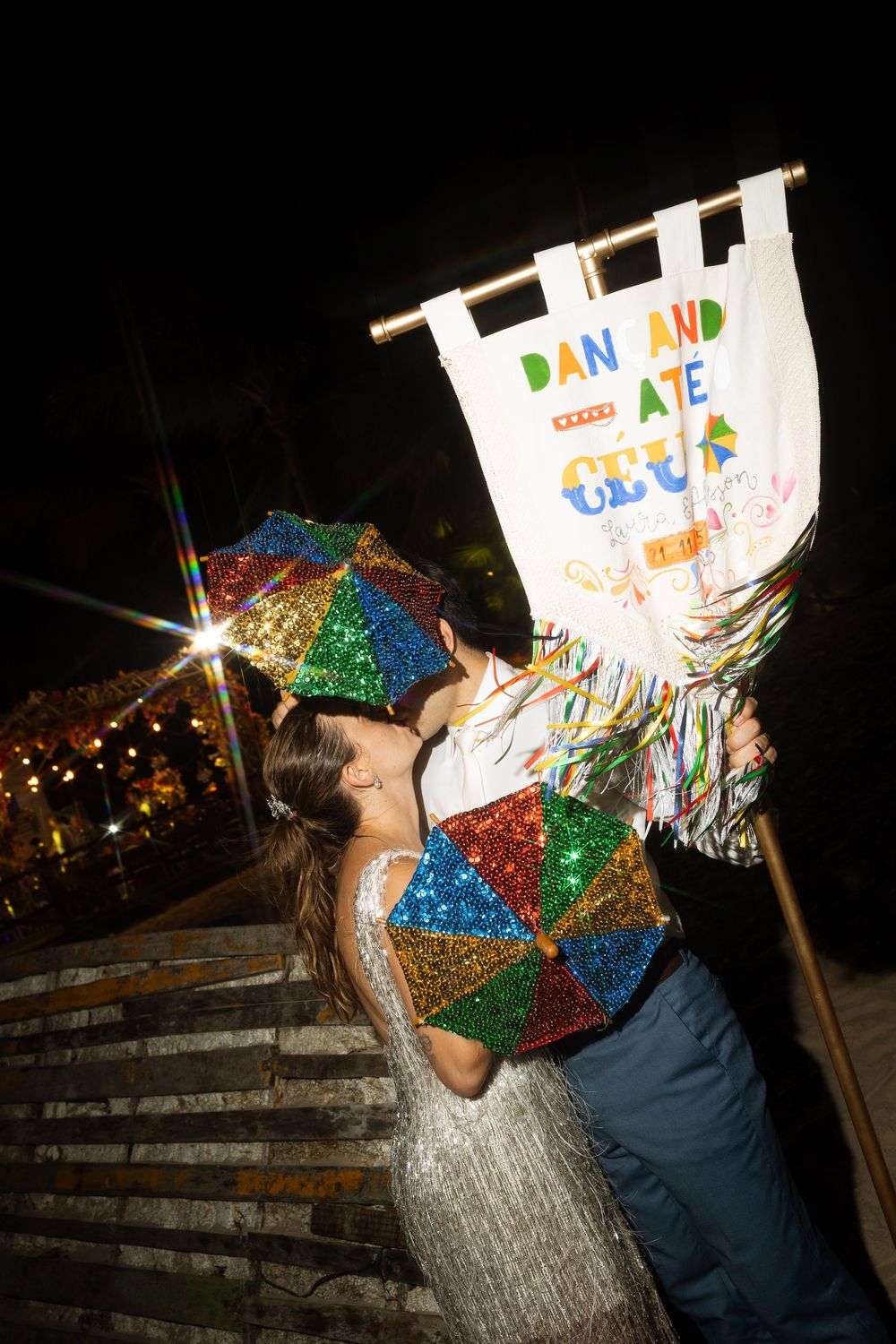 Couple kissing at night holding colorful glitter umbrellas near a festive carnival banner.