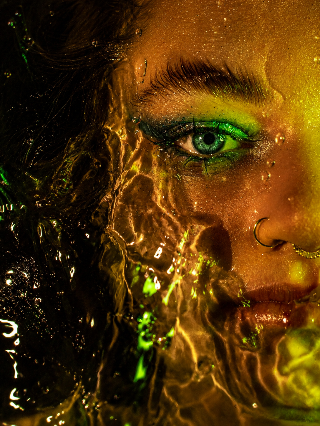 Close-up portrait of a woman with green eyeshadow and nose ring, face partially covered by golden water splash.