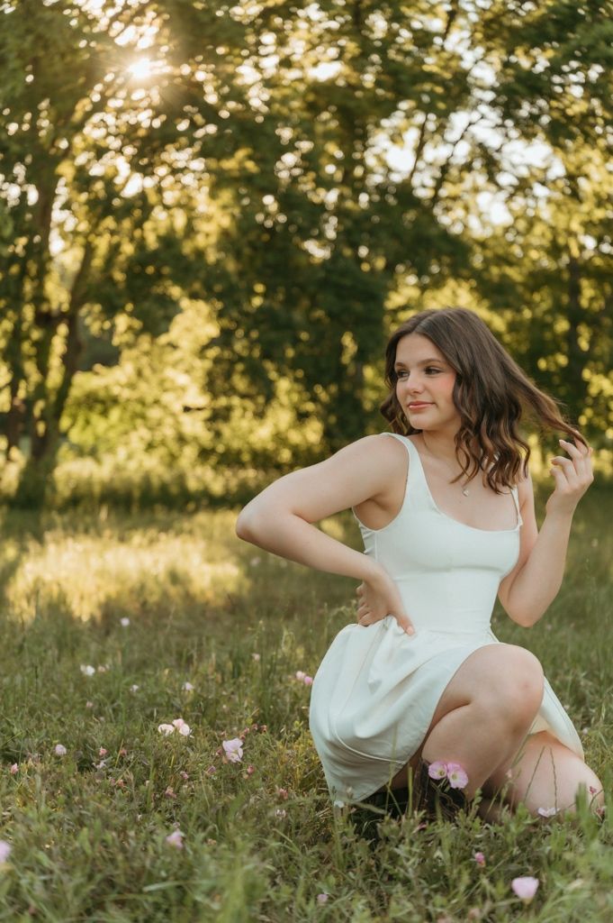 Young woman in white dress sitting gracefully in a sunlit meadow with pink wildflowers and lush green trees.