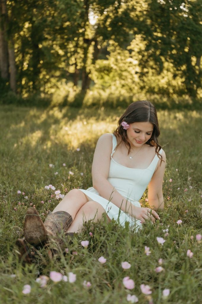 Young woman in white dress and cowboy boots sitting peacefully in a flower-filled meadow at golden hour.