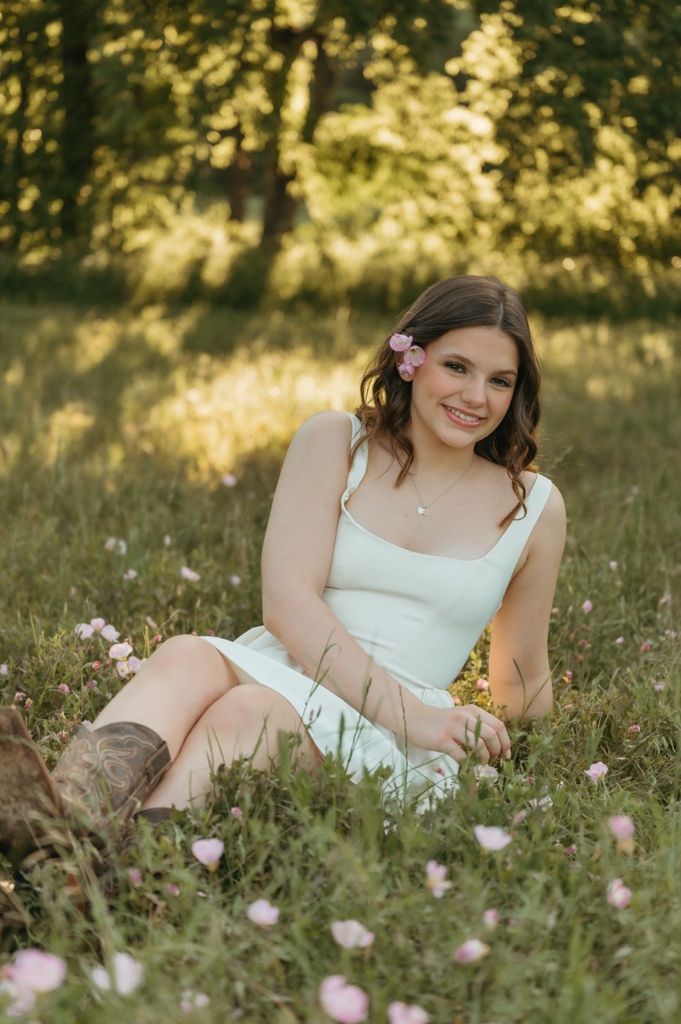 Young woman in white dress and cowboy boots sitting in a wildflower meadow with pink flowers in her hair.