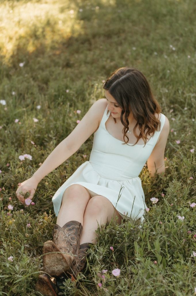 Young woman in white dress and cowboy boots sitting in a field of pink wildflowers, looking down gracefully.