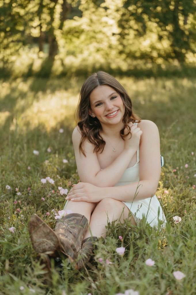 Smiling young woman in white dress and cowboy boots posing in a sunlit field of pink wildflowers.