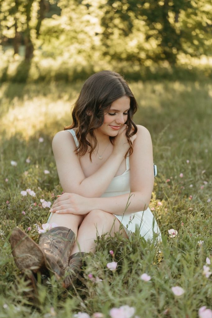 Young woman in white dress and cowboy boots sitting in a field of wildflowers, smiling softly.