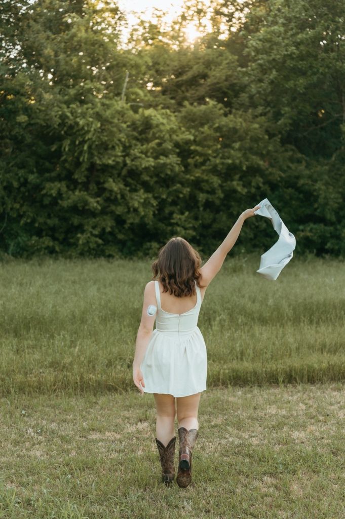 Woman in white dress and cowboy boots walks through green field at golden hour, holding fabric aloft.