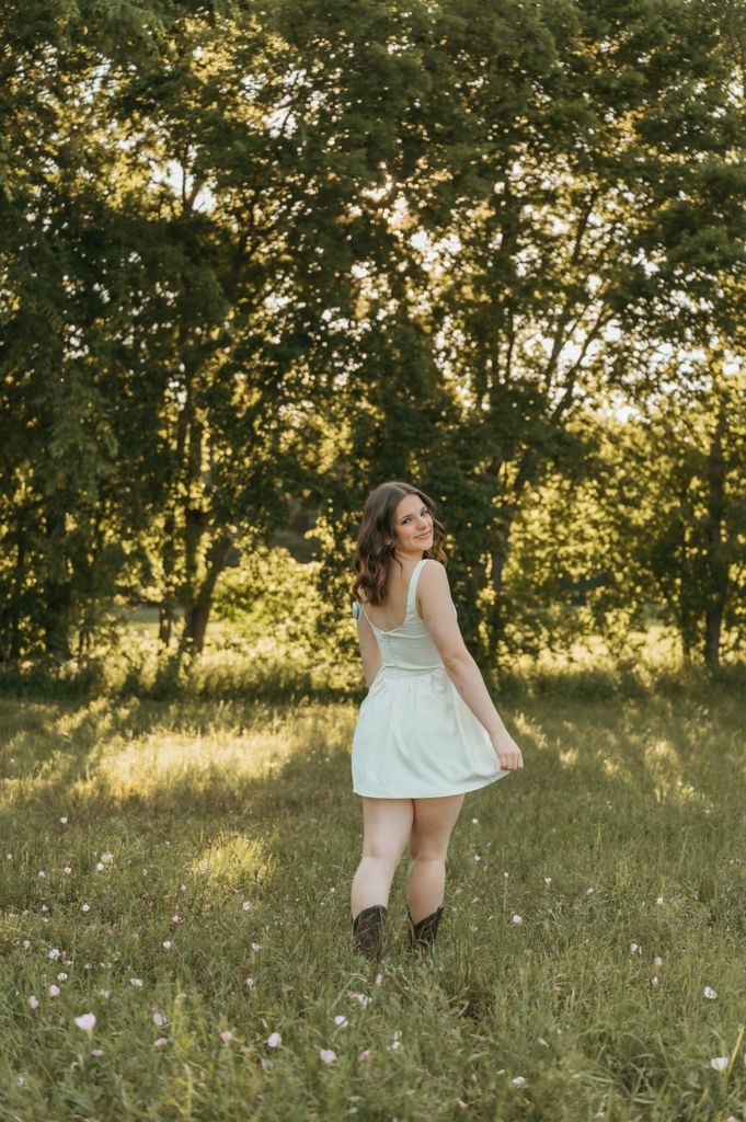 Young woman in white dress and cowboy boots twirling in a sunlit meadow surrounded by lush green trees.