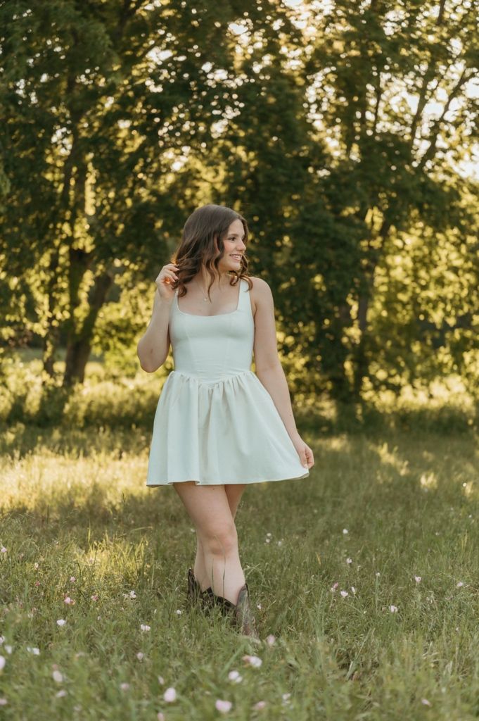 Young woman in mint green mini dress and boots standing in a sunlit meadow with lush green trees.