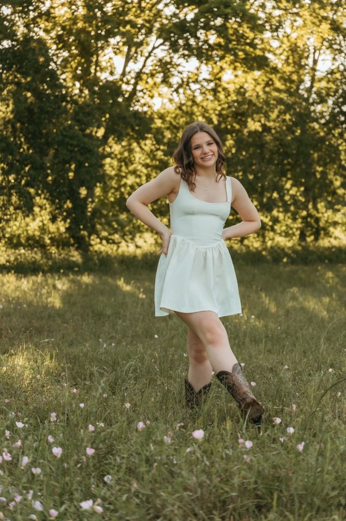 Young woman in white mini dress and cowboy boots posing in a sunny green meadow with wildflowers.