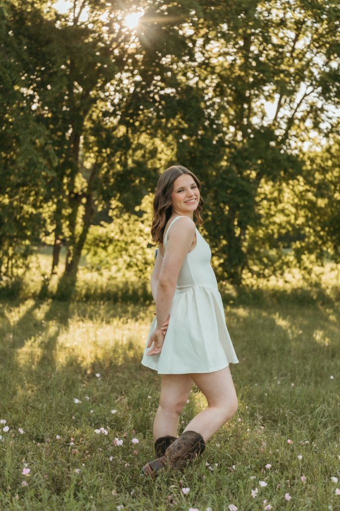 Young woman in white sundress and brown boots smiling in sunlit green meadow with trees in background.