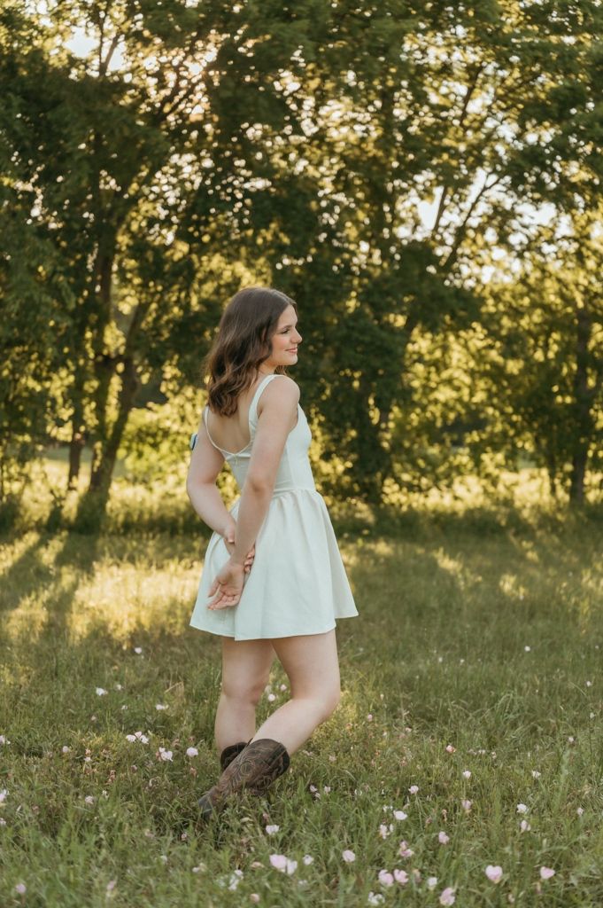Young woman in mint dress and cowboy boots standing in sunlit meadow with wildflowers and green trees.