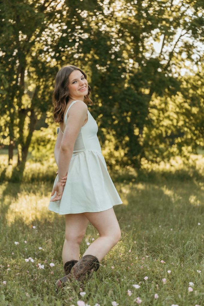 Young woman in mint dress and brown boots smiling over shoulder in sunny green meadow with wildflowers.
