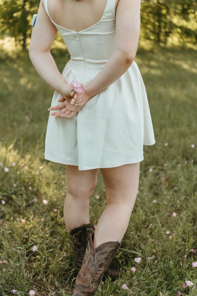 Woman in white sundress and brown cowboy boots standing in green field holding pink flowers behind her back.