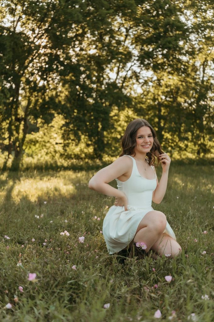 Young woman in white dress sitting gracefully in a sunlit meadow with wildflowers and lush green trees.