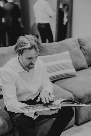 Man in white shirt reads a magazine seated on a grey sofa in black and white photo.