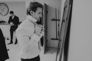 A groom adjusts his tie while getting ready in a mirror, with a groomsman visible in the background.