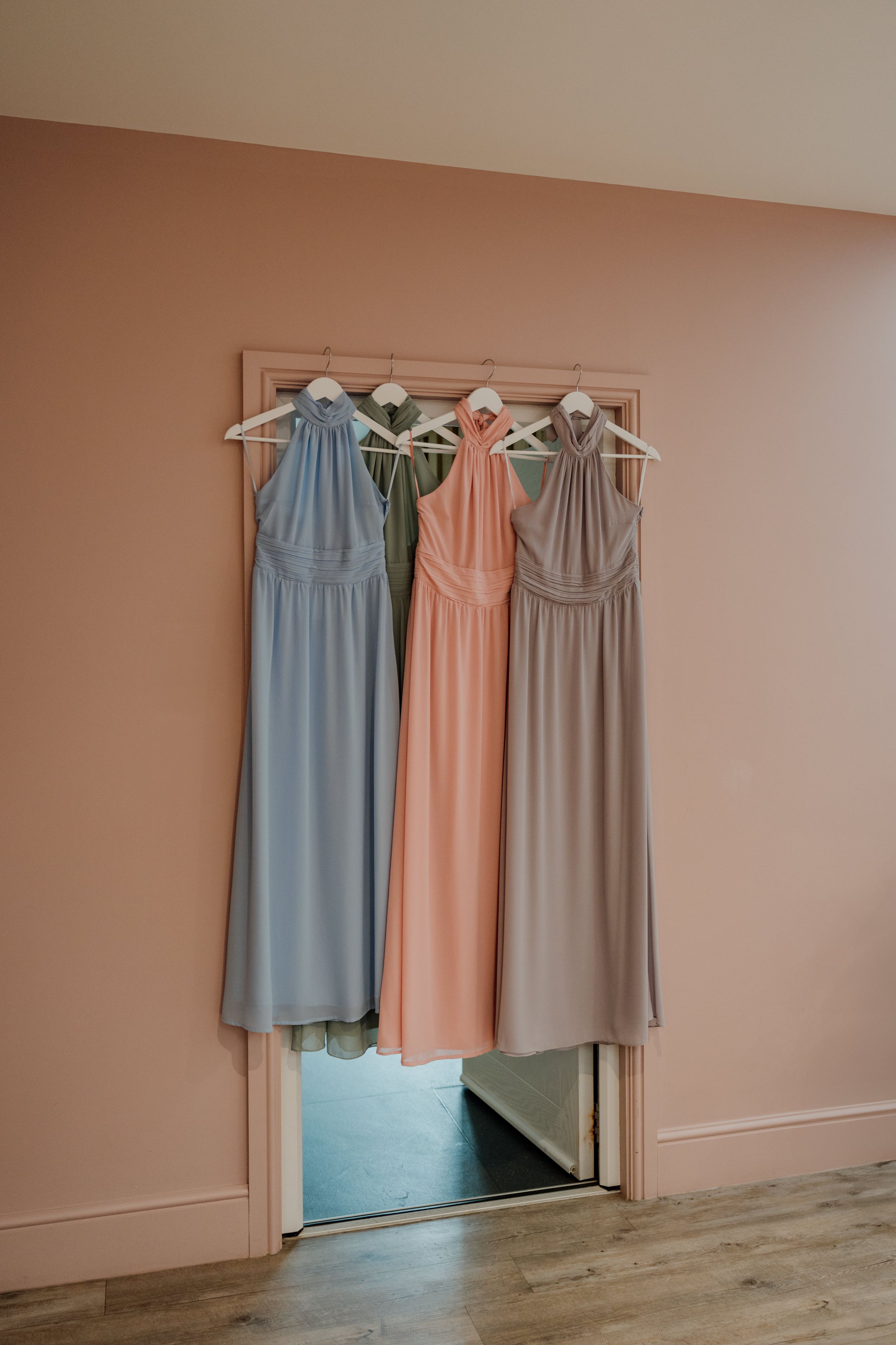 Three bridesmaid dresses in blue, pink, and taupe hanging on a mirror in a pink-walled room.