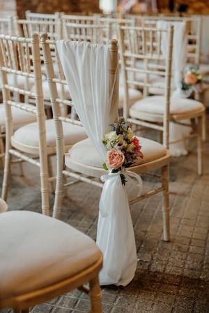 Elegant chiavari chairs with white fabric sashes and floral bouquets decorating a wedding ceremony aisle.
