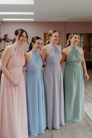 Four bridesmaids laughing in pastel halter neck gowns pink blue lavender and sage green at a wedding.