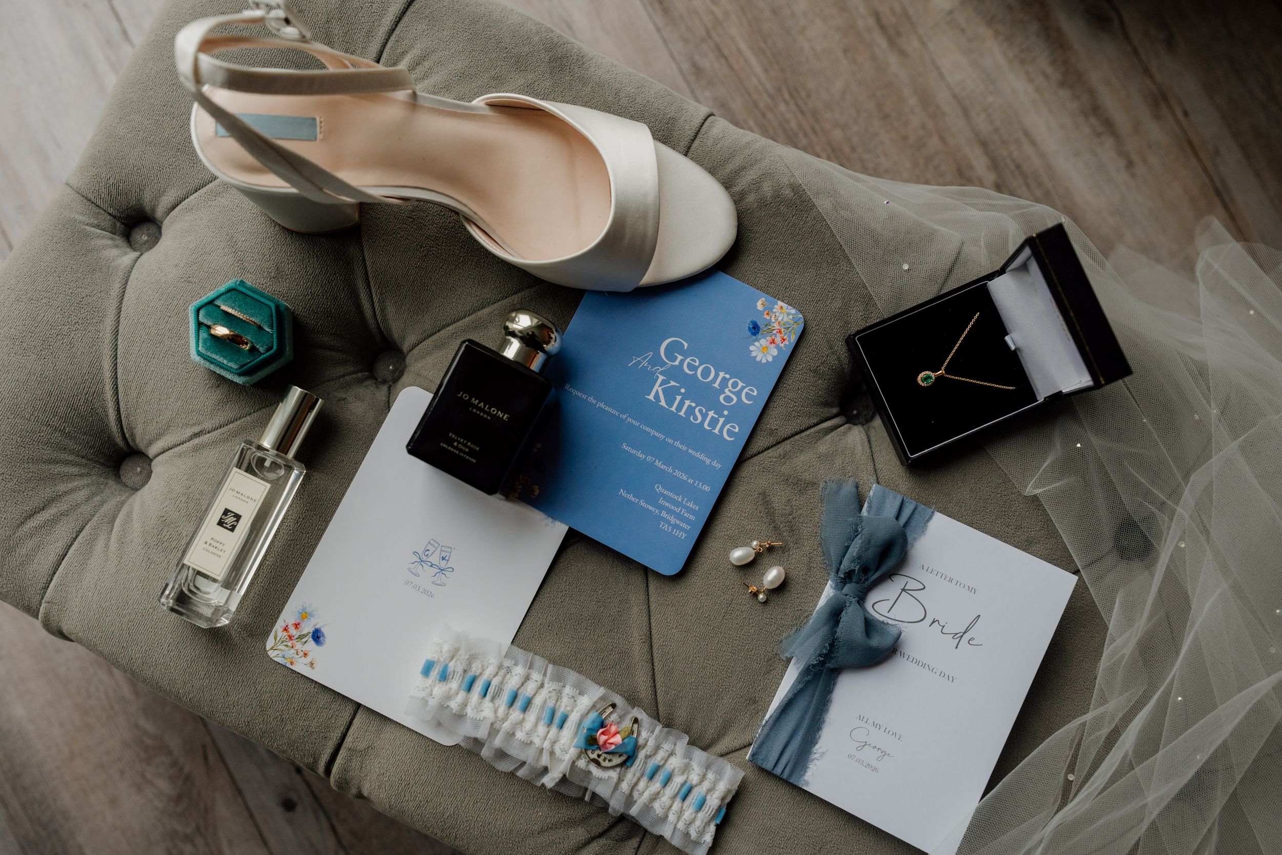 Elegant wedding flat lay featuring bridal heels, blue invitation, perfume, jewelry, and accessories on a tufted gray chair.