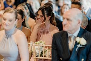 Emotional bridesmaid covering her mouth while crying during a wedding ceremony, seated among guests in elegant attire.