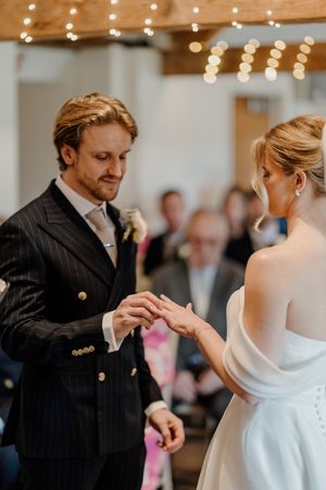Groom in pinstripe suit placing wedding ring on brides finger during indoor ceremony with fairy lights.