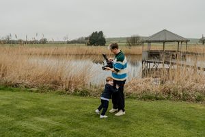 Adult and child stand by a pond with tall reeds and a gazebo on a cloudy day.