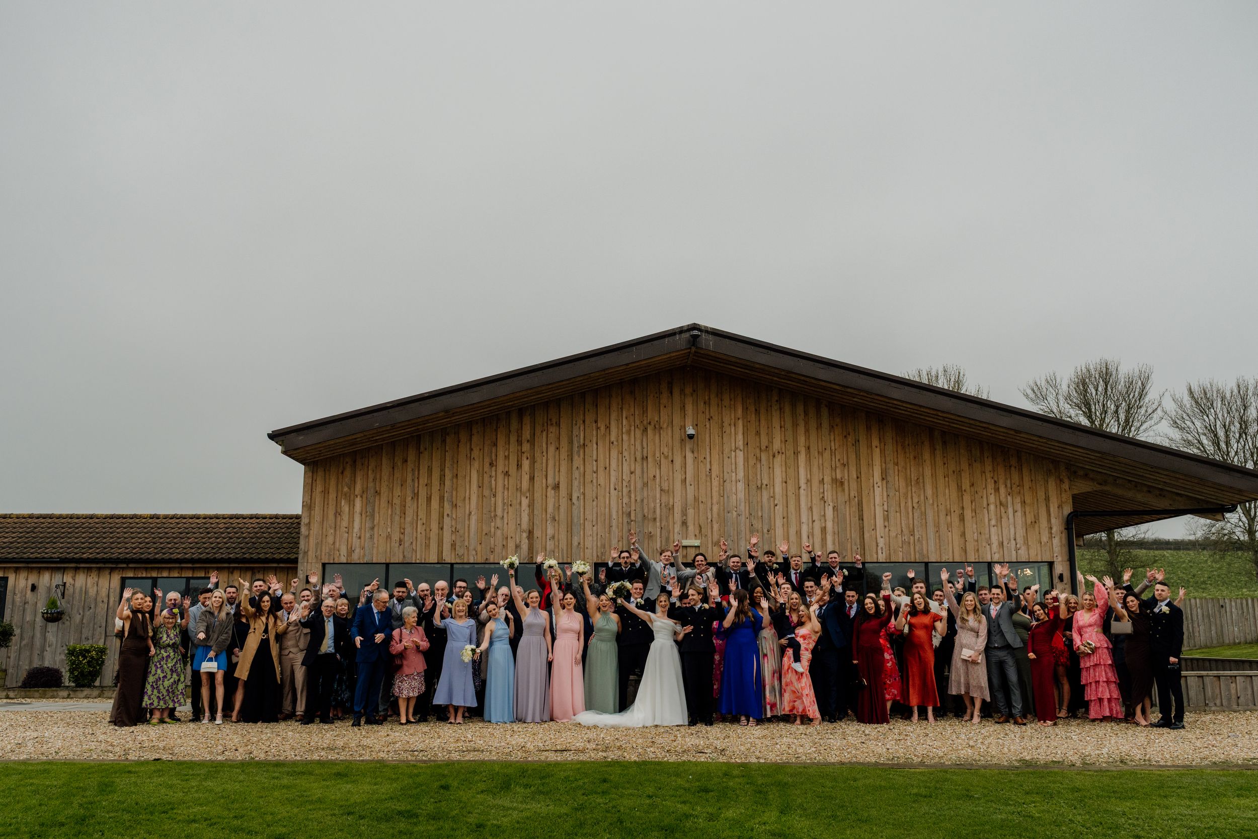 Large wedding party posing together outside a rustic wooden barn venue on an overcast day.