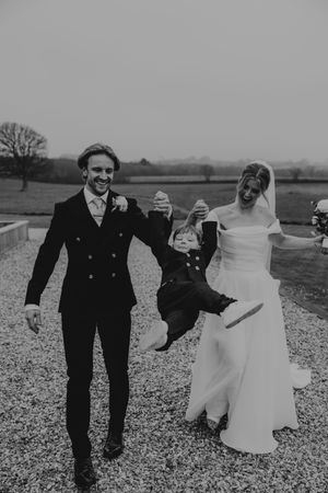 Bride and groom swing a young child between them on a gravel path at their wedding.