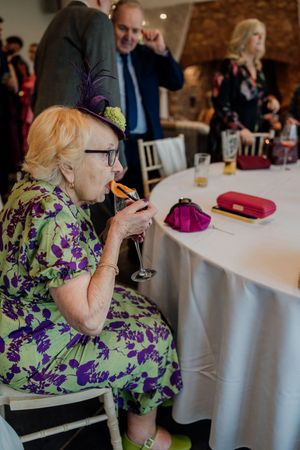 Elderly wedding guest in green floral dress sipping champagne at reception table.