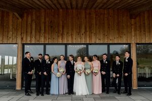 Wedding party poses outside wooden venue with bridesmaids in pastel dresses and groomsmen in dark suits.