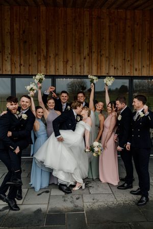 Bride and groom celebrate with their wedding party outside a modern barn venue, raising bouquets in the air.