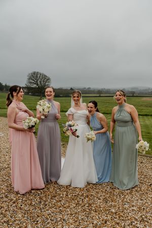 Bride poses with four bridesmaids in mismatched pastel gowns holding white bouquets on a gravel path.