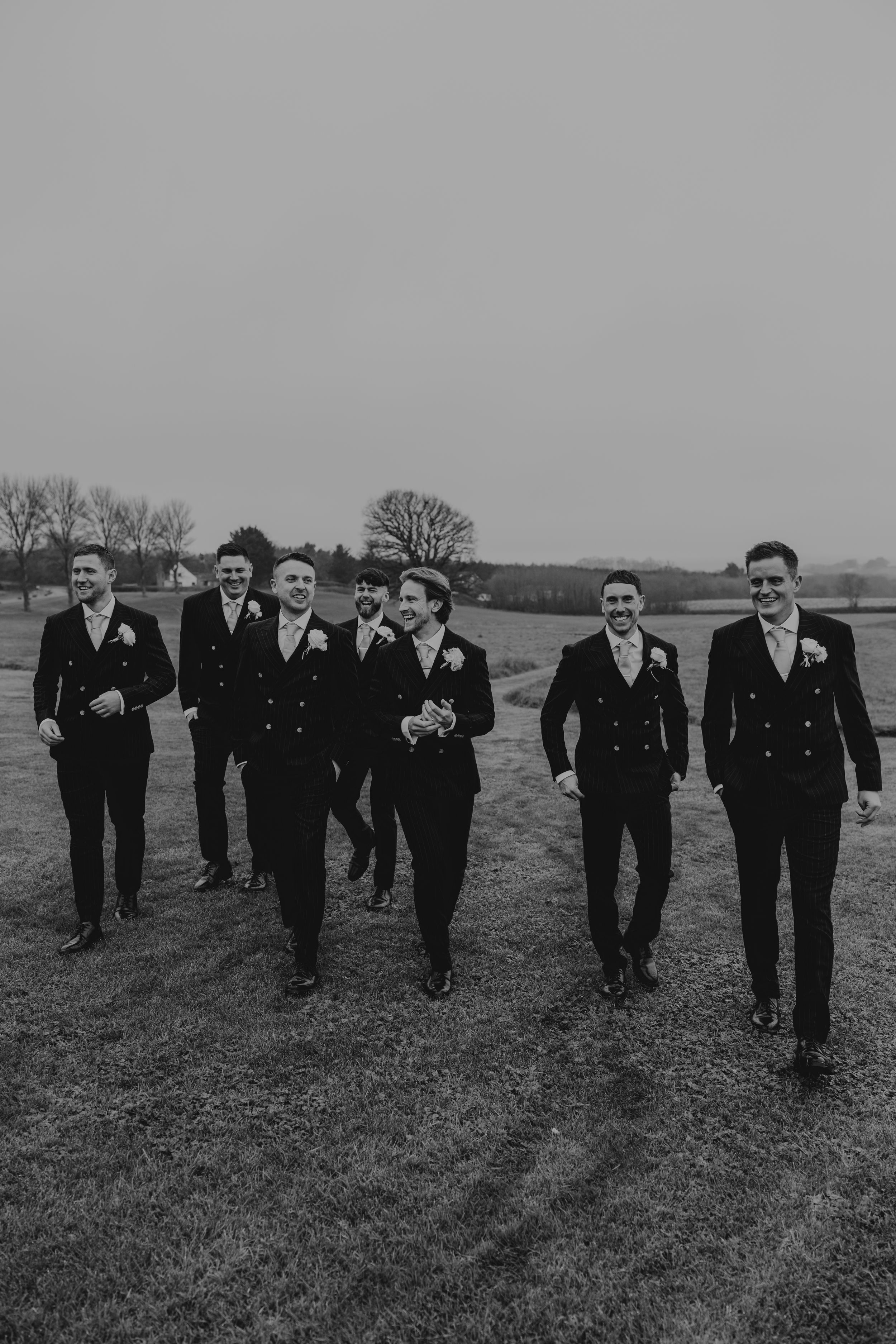 Groom and groomsmen in black suits walking across a field in black and white wedding photo.