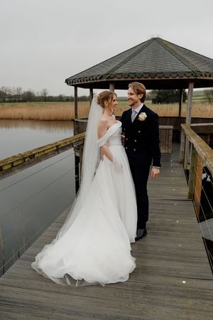 Bride and groom walking on a wooden dock by a lake, bride in an off-shoulder gown with veil, groom in navy suit.