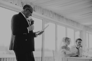 Man in suit giving a speech at a wedding reception holding microphone and notes in black and white photo.