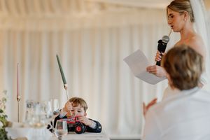 Bride giving speech at wedding reception while young child plays at table.