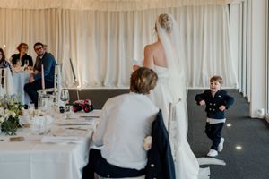Bride in white gown and veil greeting a young boy running toward her at an elegant wedding reception.