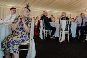 Elderly woman in purple floral dress sitting at a wedding reception table with hands clasped together.