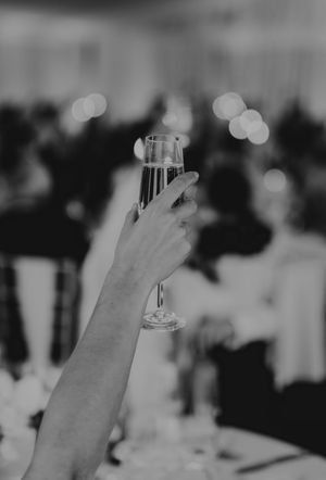 Hand raises a champagne flute in a toast at a wedding reception in black and white.