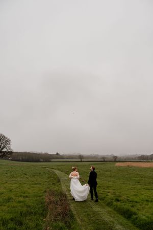 Bride and groom walk hand in hand through a misty green countryside field on their wedding day.