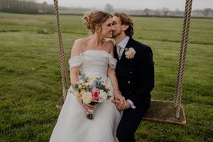 Bride and groom sharing a kiss on a rope swing in a green field, bride holding a pastel bouquet in off-shoulder gown.