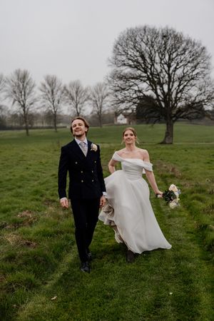 Bride and groom walk hand in hand across a green field with bare trees behind them.