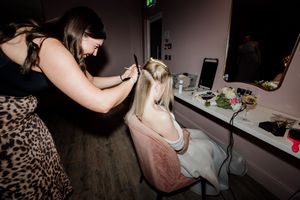A makeup artist applies makeup to a blonde woman seated at a vanity mirror in a salon.