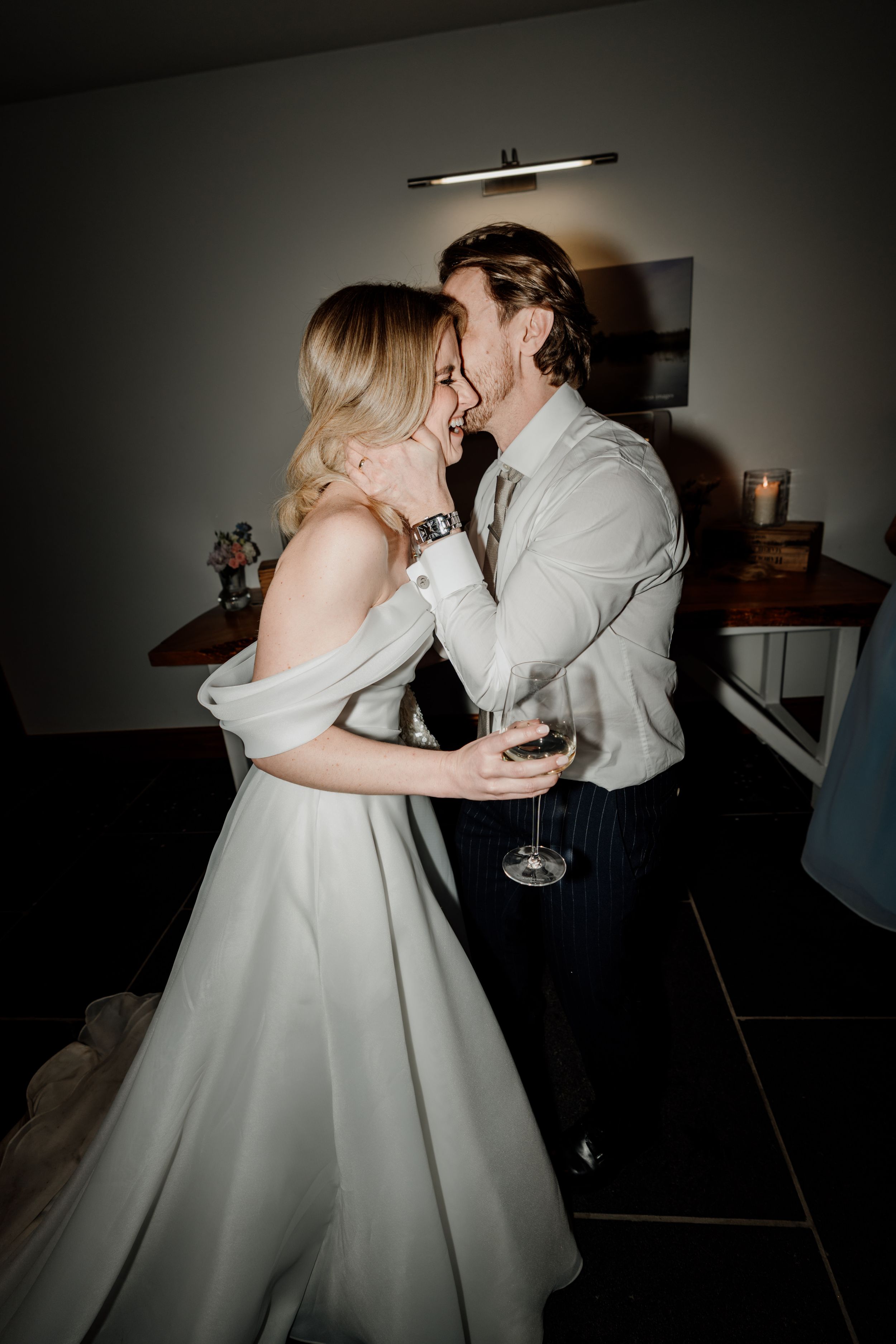 A bride in a white off-shoulder gown and groom in a white shirt share a romantic embrace, holding champagne indoors.