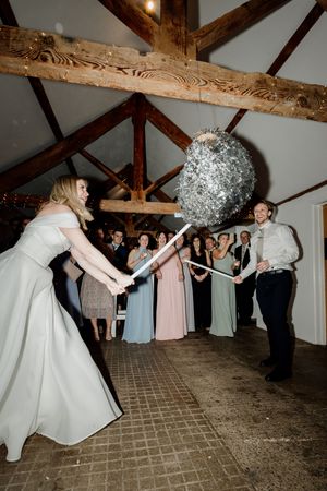 A bride and groom joyfully smash a silver glitter pinata with sticks at their rustic barn wedding reception.