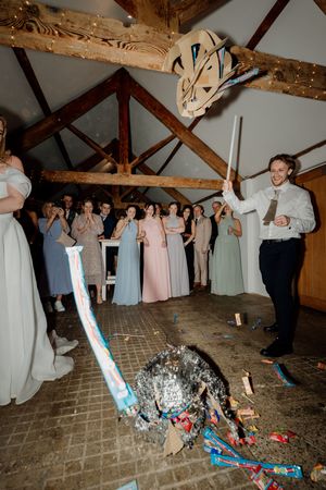 Groom smashing a pinata at wedding reception in rustic barn venue with guests watching and cheering.