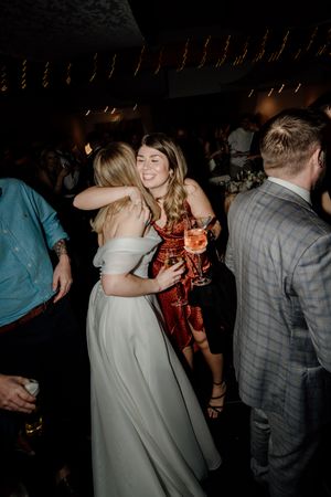 Bride in white gown hugs a guest in a red dress at her wedding reception.