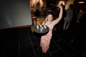 Woman in floral dress carrying tray of shot glasses at wedding reception bar area.