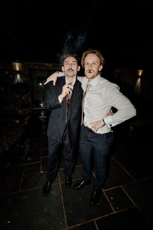 Two men in formal suits pose together outdoors at night, smoking cigars at a wedding celebration.