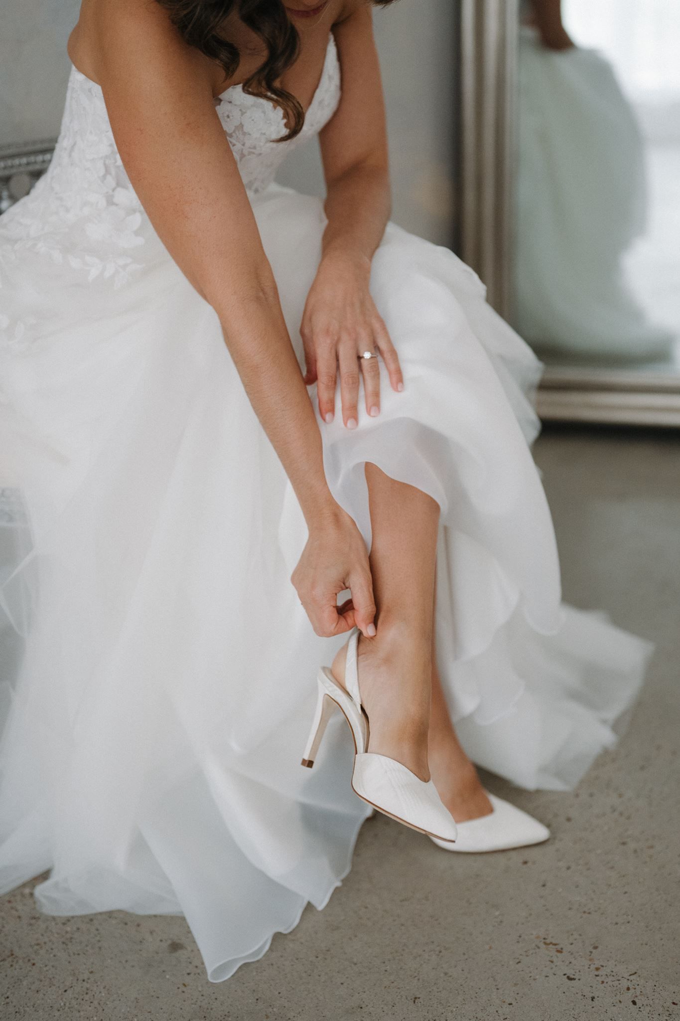 Bride in white wedding gown putting on ivory satin stiletto heels, wearing engagement ring, seated before mirror.